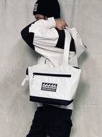 Person wearing a white jacket and black pants with a white tote bag featuring a logo on a gray background