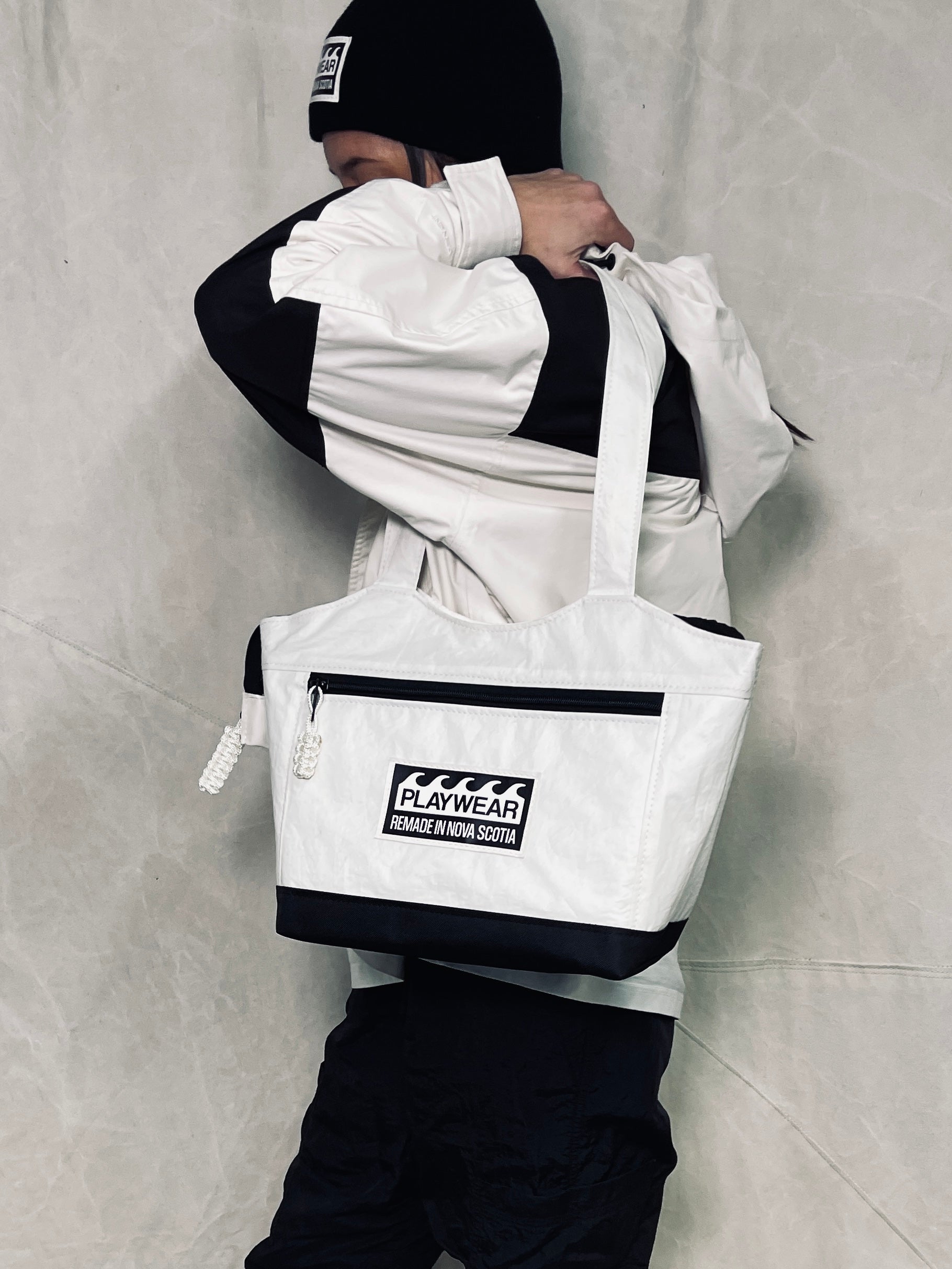 Person wearing a white jacket and black pants with a white tote bag featuring a logo on a gray background