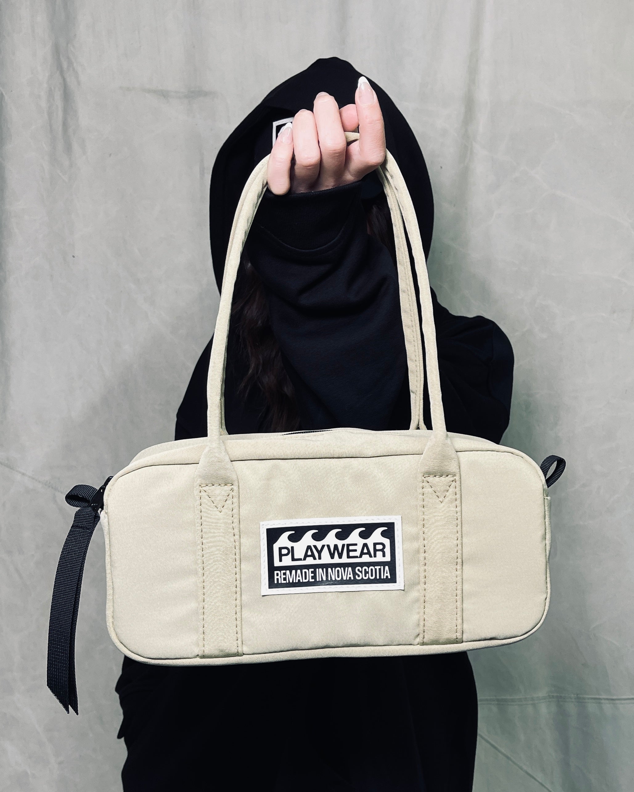 Beige handbag with a visible brand label held by a person wearing a black outfit against a gray background