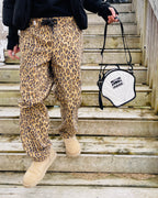 Person wearing leopard print pants and beige boots holding a white bag with black strap on wooden stairs.