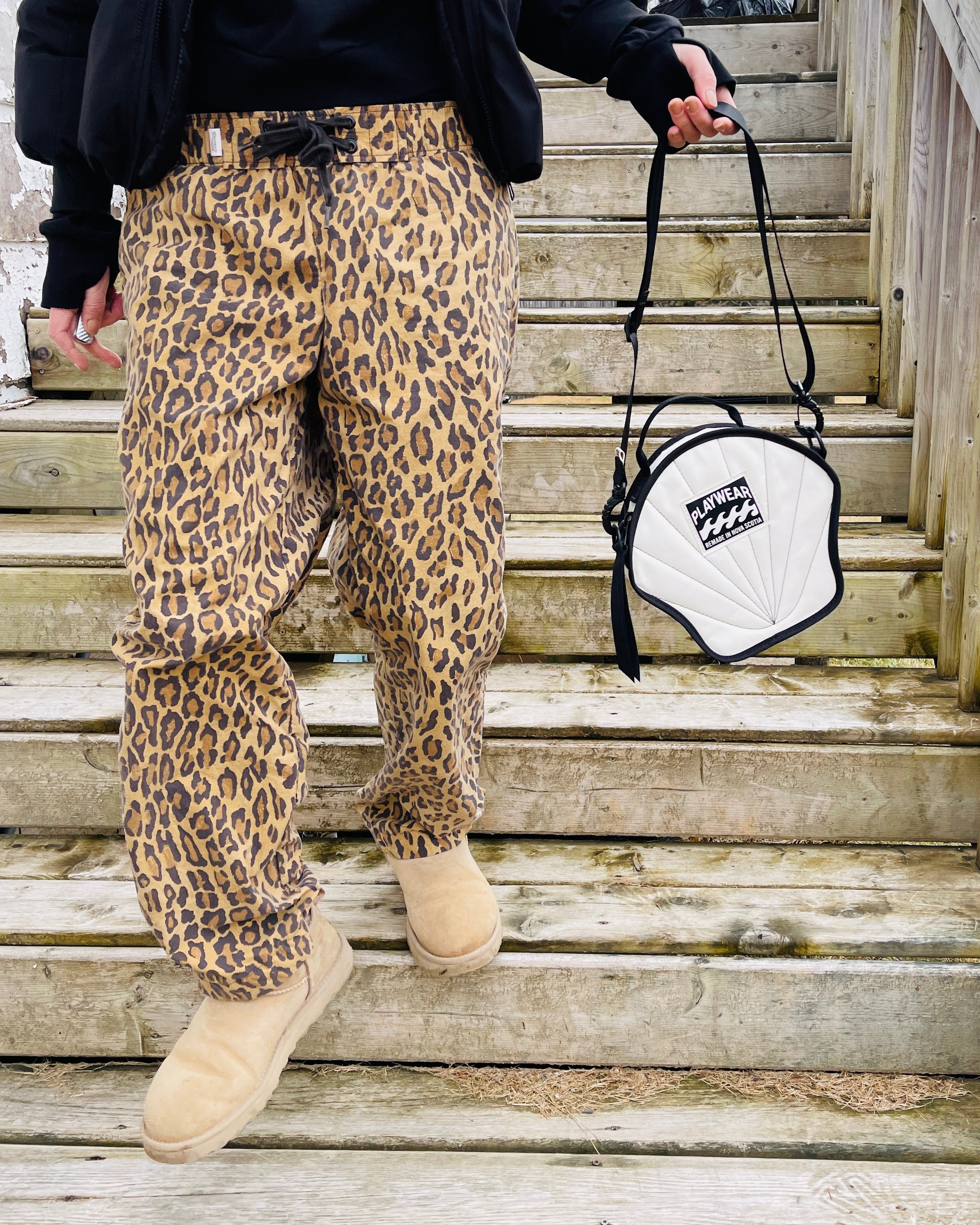 Person wearing leopard print pants and beige boots holding a white bag with black strap on wooden stairs.