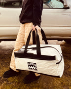 Person holding a white duffel bag with black handles and a visible brand logo, standing next to a car. MADE IN NOVA SCOTIA CANADA.