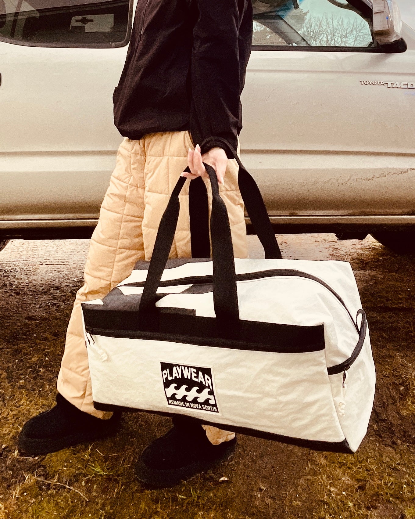 Person holding a white duffel bag with black handles and a visible brand logo, standing next to a car. MADE IN NOVA SCOTIA CANADA.